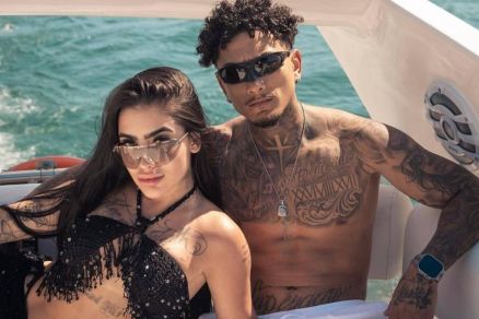 Um casal est posando em um barco, com o mar ao fundo. A mulher est usando um biquni preto com detalhes brilhantes e culos escuros, enquanto o homem est sem camisa, usando shorts brancos e tambm culos escuros. Ambos tm tatuagens visveis e esto sor