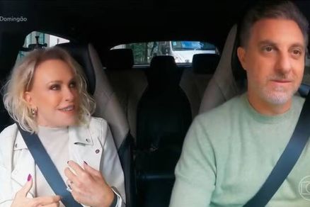 A imagem mostra duas pessoas dentro de um carro.  esquerda, uma mulher com cabelo loiro e curto, usando uma jaqueta branca, parece estar conversando animadamente.  direita, um homem com cabelo curto e grisalho, vestido com um suter verde claro, est ouv