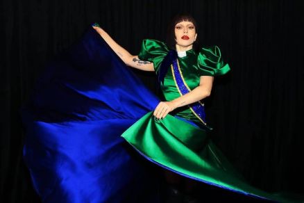 Uma modelo est posando em um fundo preto, vestindo um vestido de gala com mangas volumosas. O vestido  predominantemente verde com detalhes em azul, e a modelo est segurando uma parte do vestido, fazendo com que ele se espalhe. Ela tem cabelo curto e us