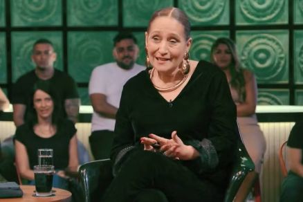 Uma mulher de cabelos presos, vestindo uma blusa preta, est sentada em uma cadeira, sorrindo e gesticulando enquanto fala. Ela usa brincos grandes e colares. Ao fundo, h um grupo de pessoas assistindo, algumas delas sorrindo. A mesa  sua frente tem um c