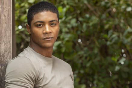Um homem est posando em um ambiente natural, encostado em uma estrutura de madeira. Ele usa uma camiseta de cor clara e calas escuras. O fundo  composto por folhagens verdes, criando um contraste com a roupa do homem. Ele tem uma expresso confiante e e