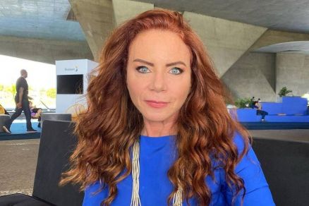 A imagem mostra uma mulher com cabelo cacheado e ruivo, vestindo uma blusa azul. Ela est sentada em um sof preto, com um fundo que apresenta uma estrutura arquitetnica moderna. A mulher olha diretamente para a cmera, com uma expresso sria. Ao fundo, 