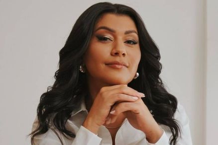 A imagem mostra uma mulher com cabelo longo e ondulado, usando uma camisa branca. Ela est sentada, com as mos unidas em frente ao rosto, e apresenta uma expresso confiante. Ao fundo, h uma planta verde, que adiciona um toque natural ao ambiente. O cen