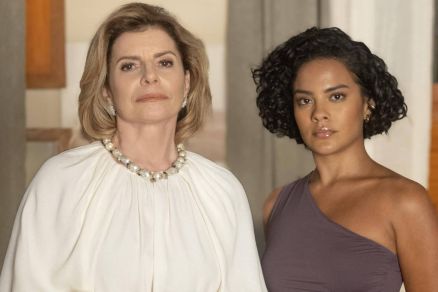 A imagem mostra duas mulheres em p, uma ao lado da outra. A mulher  esquerda tem cabelo loiro e usa uma blusa branca com um colar de prolas. A mulher  direita tem cabelo curto e cacheado, vestindo uma blusa de um ombro s na cor roxa. O fundo  compost
