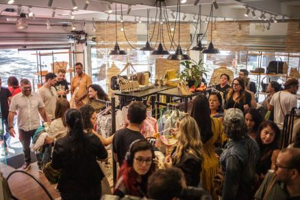 Interior de loja com muitas pessoas circulando entre araras e prateleiras de roupas e bolsas. Ambiente iluminado por luz natural e luminrias pendentes, com paredes de madeira clara e decorao moderna. Fs de Hebe Camargo lotam brech Pea Rara, no bairro